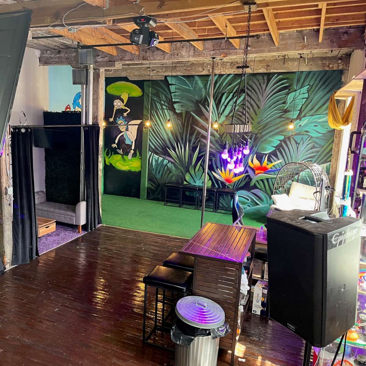 An indoor space with colorful murals, a wooden floor, seating areas, and a lush green backdrop.
