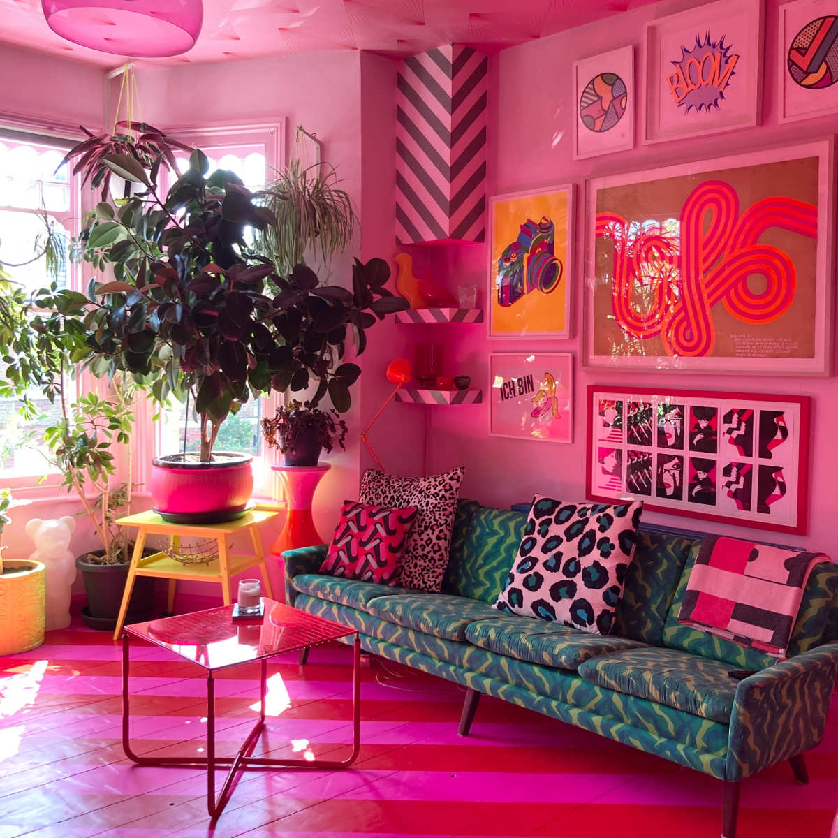 A brightly decorated living room with a pink color scheme, featuring a green sofa, various plants, and colorful artwork on the walls.