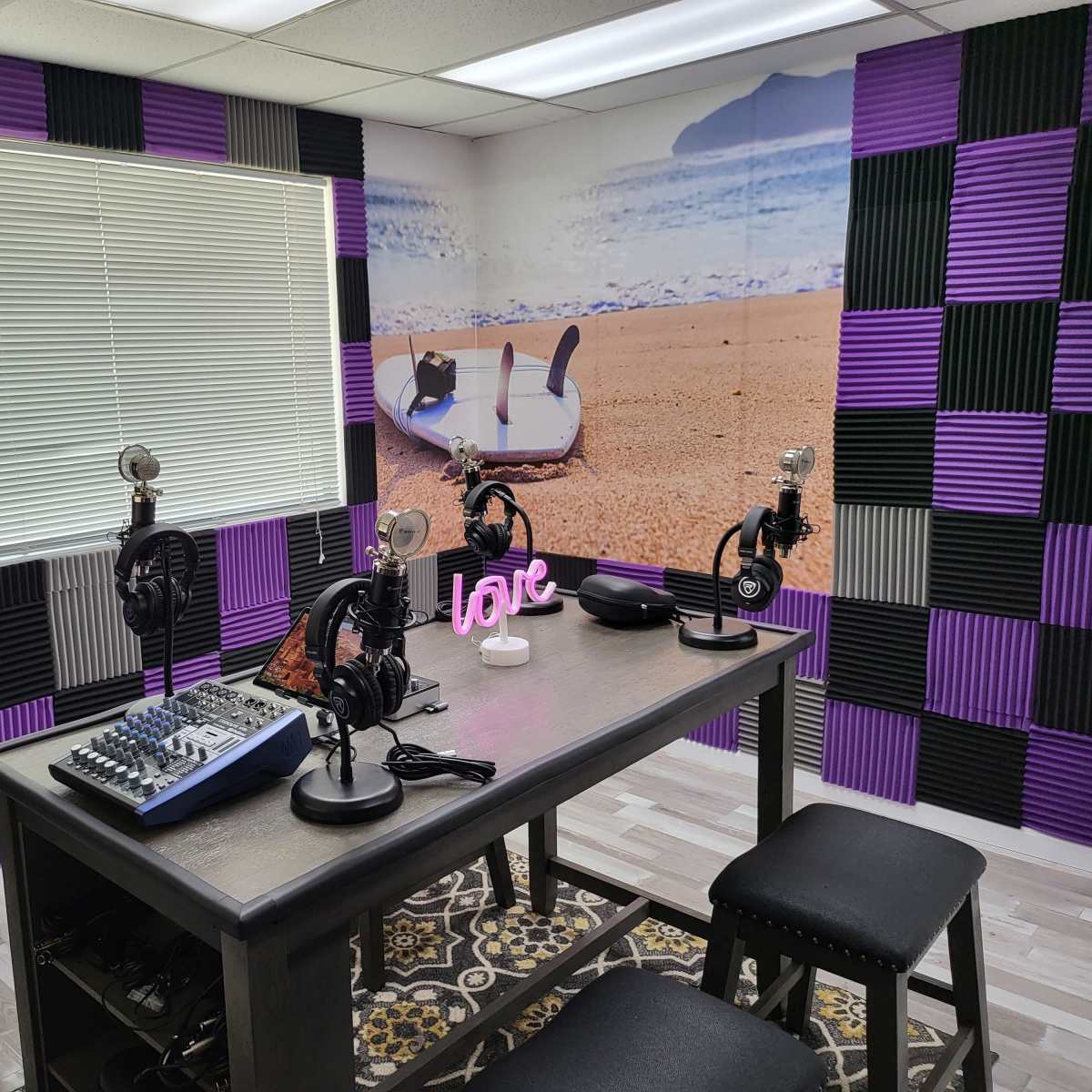 A recording studio with three microphones on stands arranged around a table, colorful soundproofing panels on the walls, and a beach-themed mural.