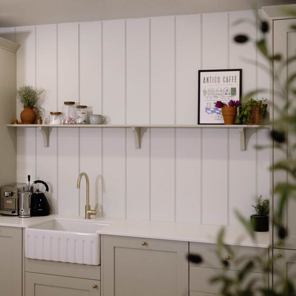 A modern kitchen featuring a classic farmhouse sink, cabinetry, and a minimalistic shelf with decorative plants and kitchenware.