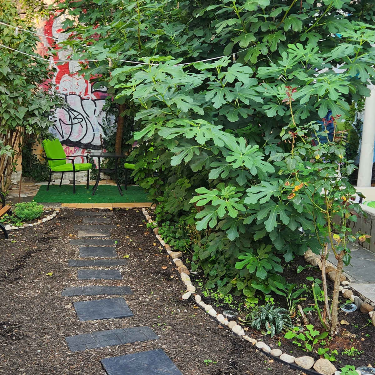A small garden with a stone path, a wooden bench, and lush greenery, bordered by a wall and a seating area.