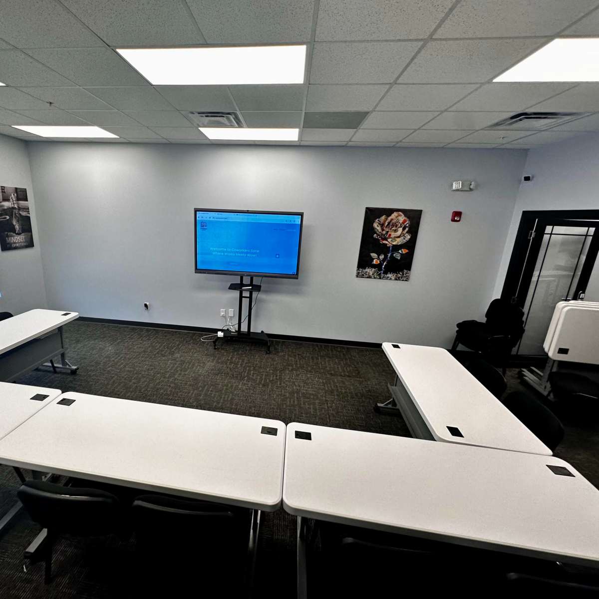 A bright, modern conference room features several tables arranged in a U-shape facing a mounted screen, with wall art on the sides.