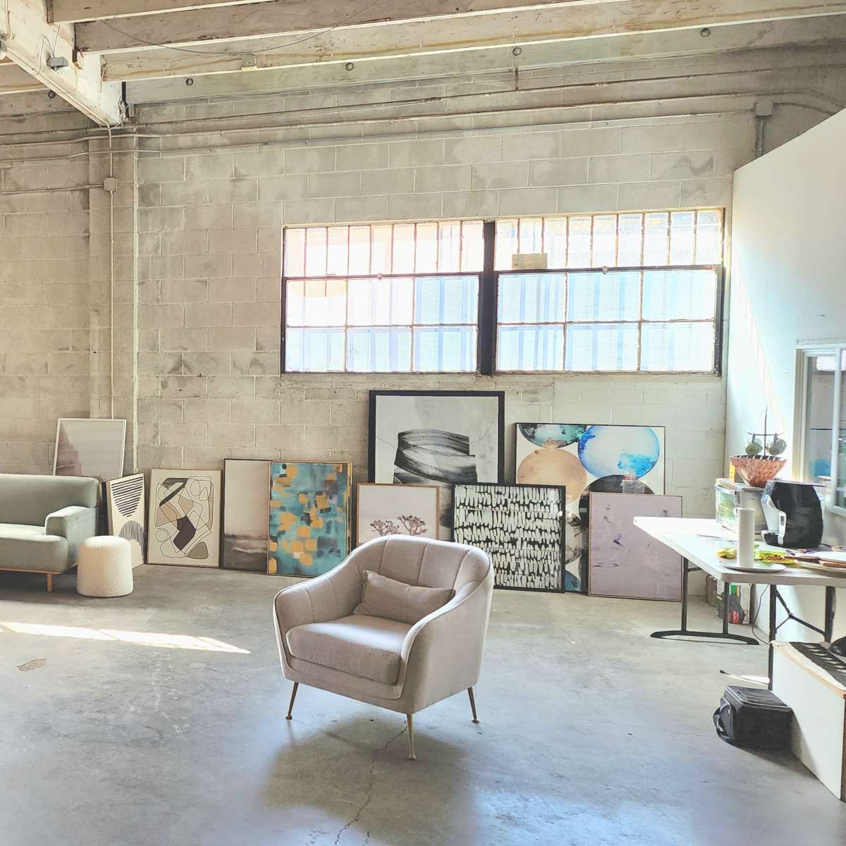 A spacious studio features a dog on the floor, a green sofa, a light gray armchair, and various artworks displayed against the walls.