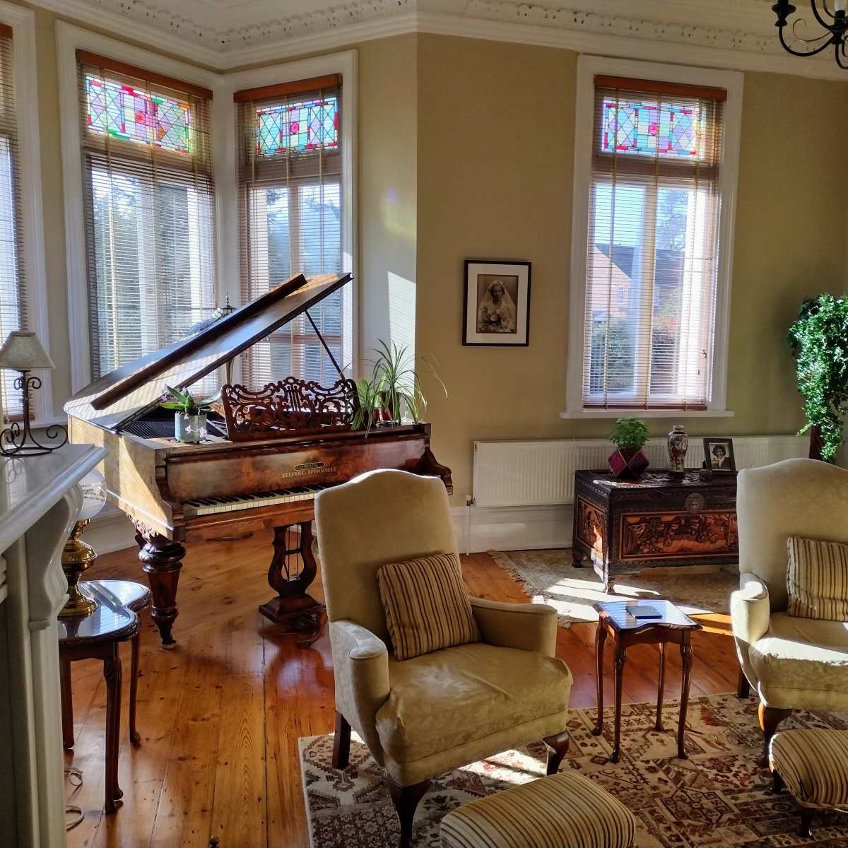 A sunlit living room featuring a piano, two upholstered chairs, a decorative rug, and stained glass windows.