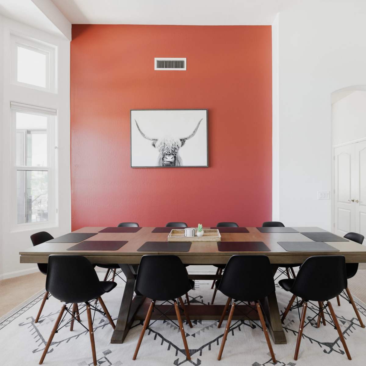 A long wooden dining table with black chairs is set against a red accent wall featuring a framed picture of a bull's head.