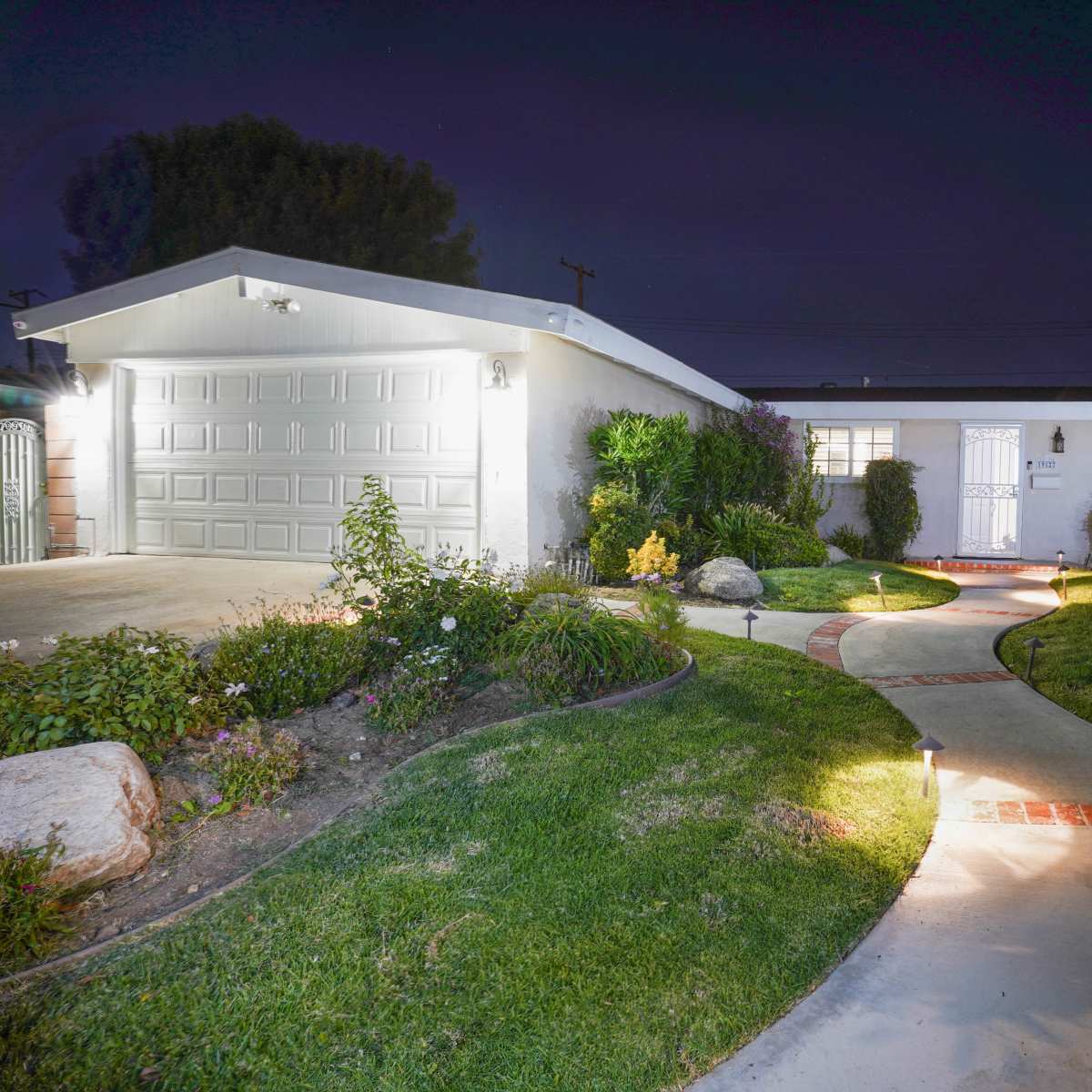 A well-lit pathway leads to a single-story home with a garage, surrounded by manicured lawns and flower beds at night.