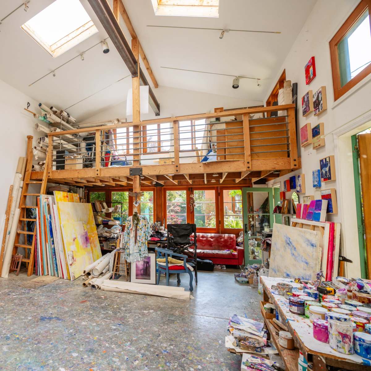 An art studio with high ceilings, large windows, and various paintings displayed on the walls and scattered around the space.