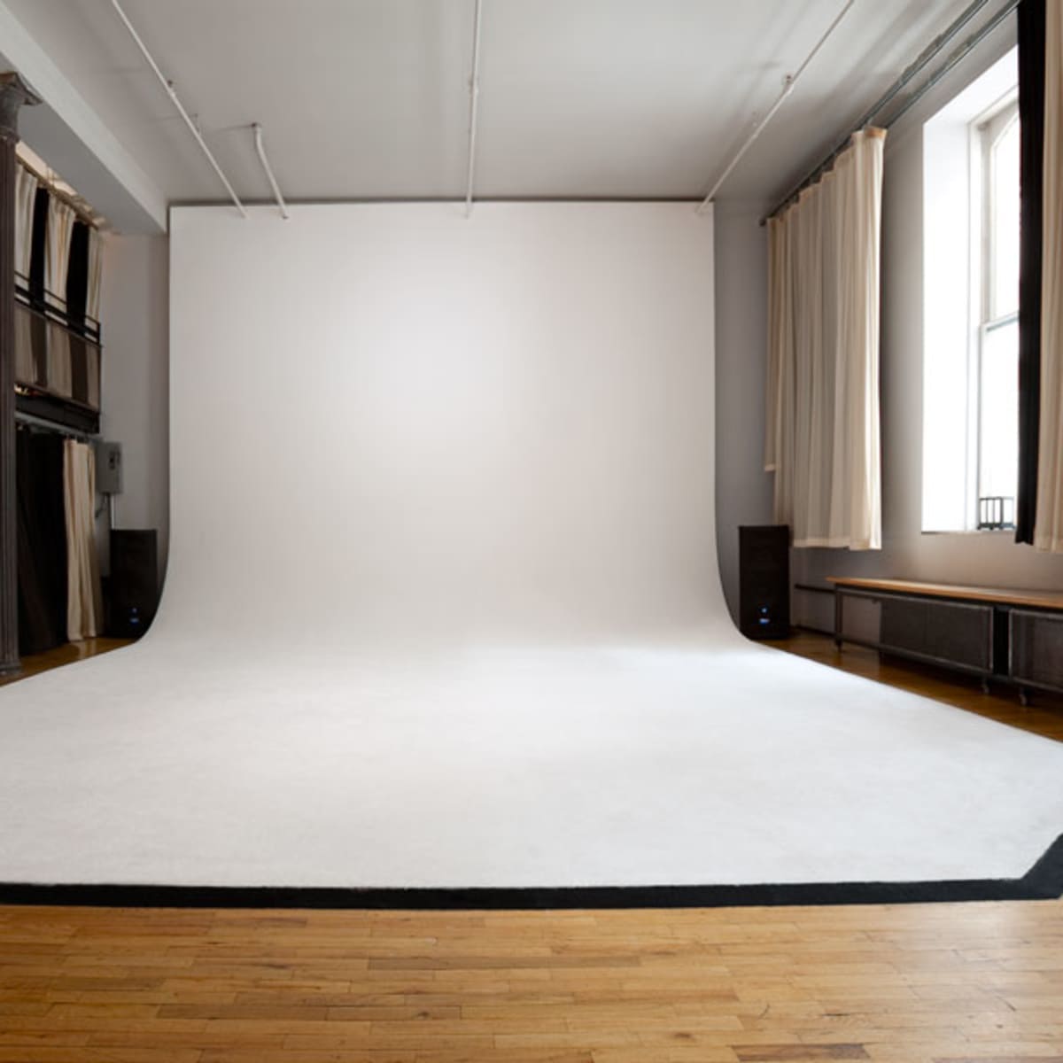 A spacious photo studio with a large, white backdrop and a wooden floor, illuminated by natural light from windows.