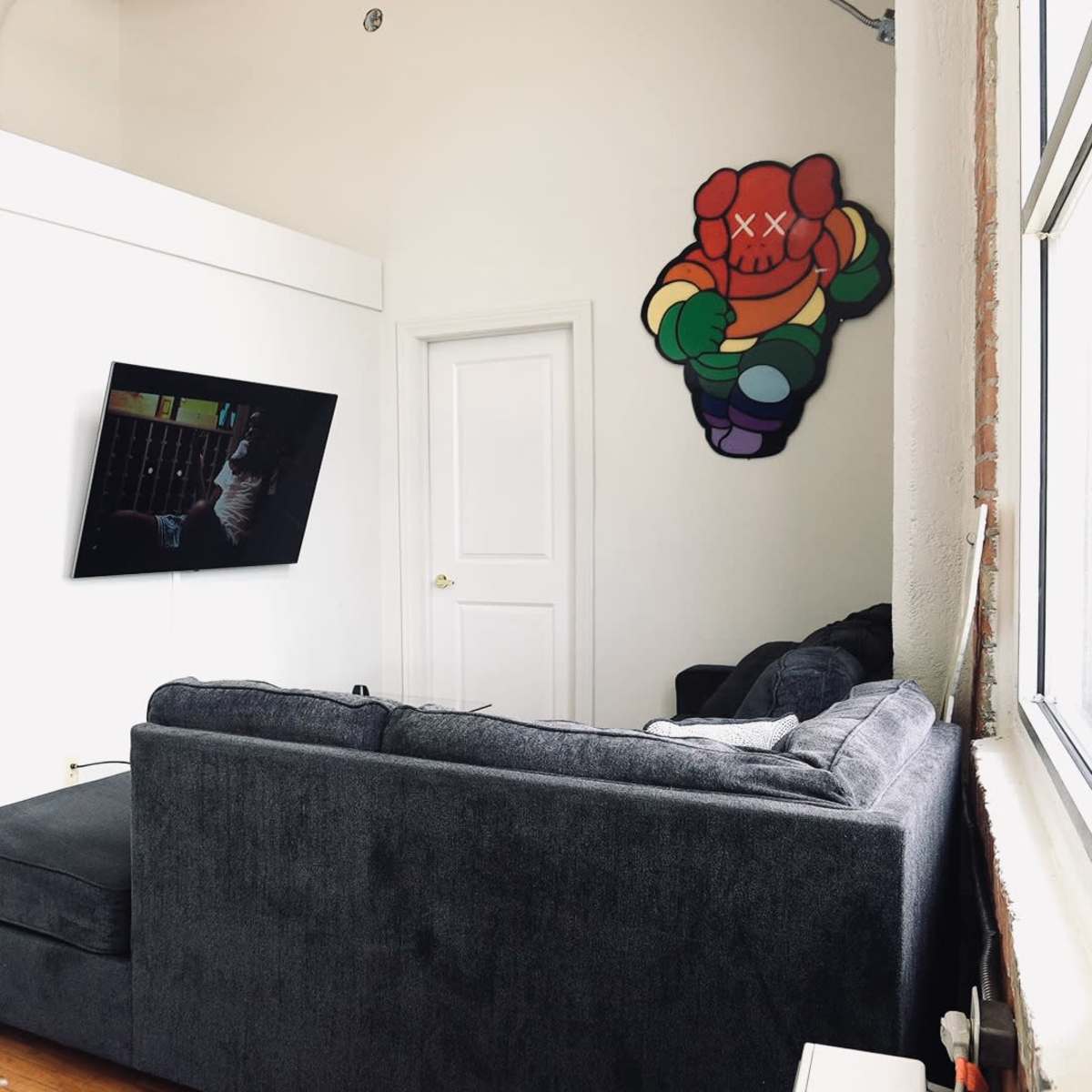 A living room with a dark gray sectional sofa, a wall-mounted television, and a colorful bear-shaped mural on the white wall.