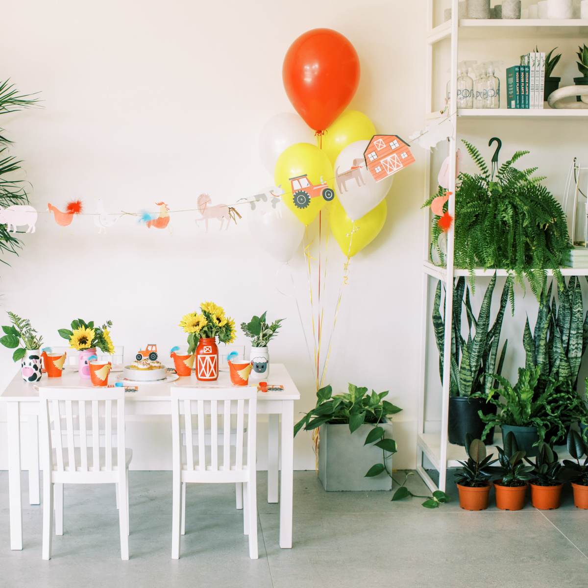 A white table is set with colorful tableware and decor for a celebration, surrounded by potted plants and balloons in a bright room.
