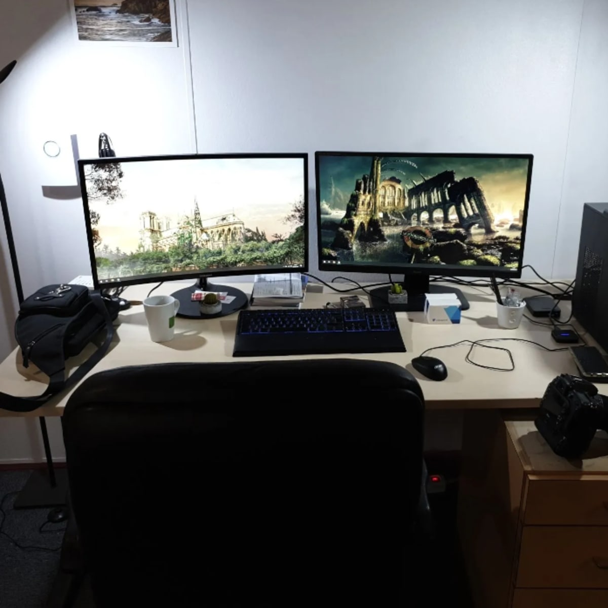 A workspace features a desk with two monitors displaying images, a gaming keyboard, a camera, and various accessories, set against a plain wall.