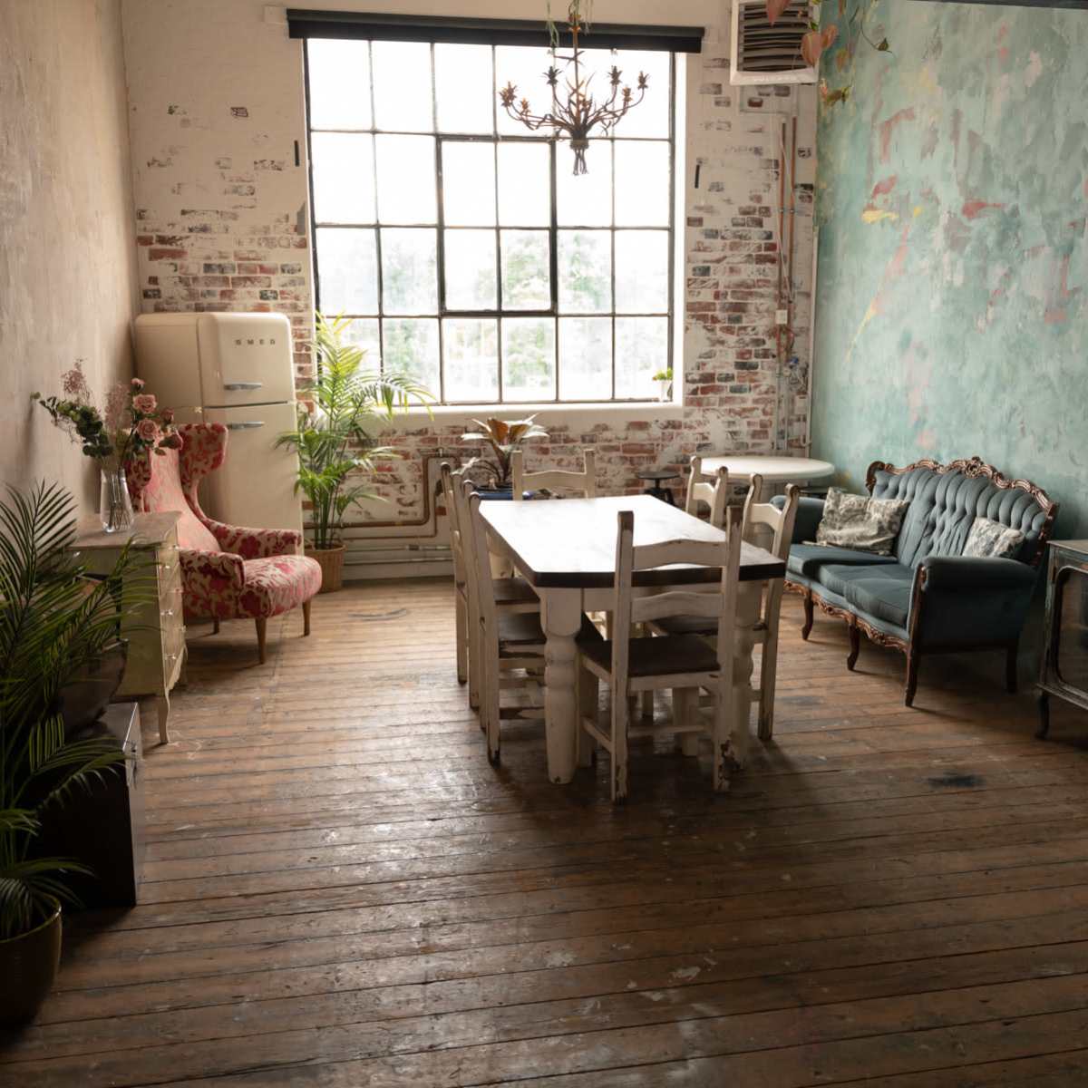 A spacious room with wooden floors, a large window, a dining table surrounded by chairs, a vintage couch, and a retro-style refrigerator.