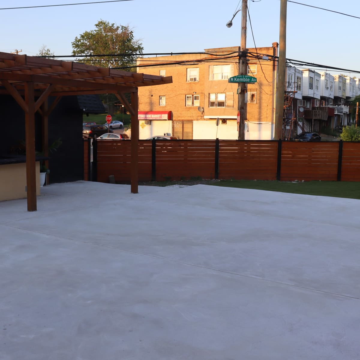 A partially finished outdoor patio area with a wooden pergola and a fence, overlooking a street with parked cars and buildings in the background.