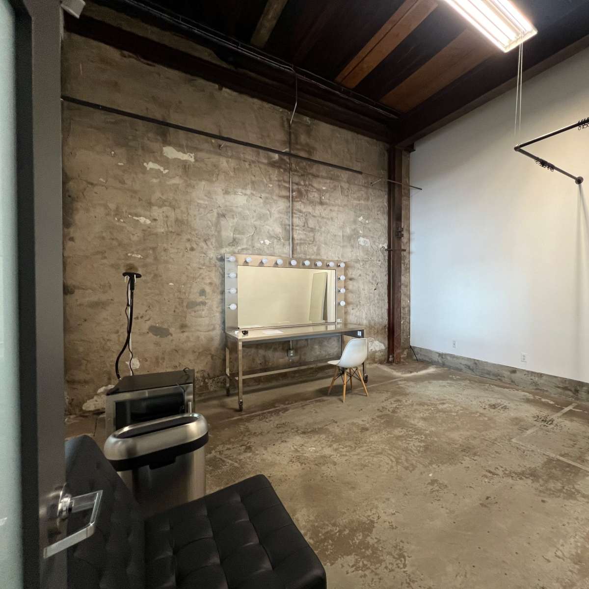 An empty salon or dressing room with a large mirror, a lighted vanity, a chair, and concrete walls and floors.