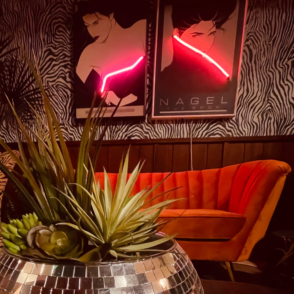 A decorative indoor arrangement features a shiny, mirrored pot with plants in the foreground, contrasting with a vibrant orange couch and illuminated art posters in the background.