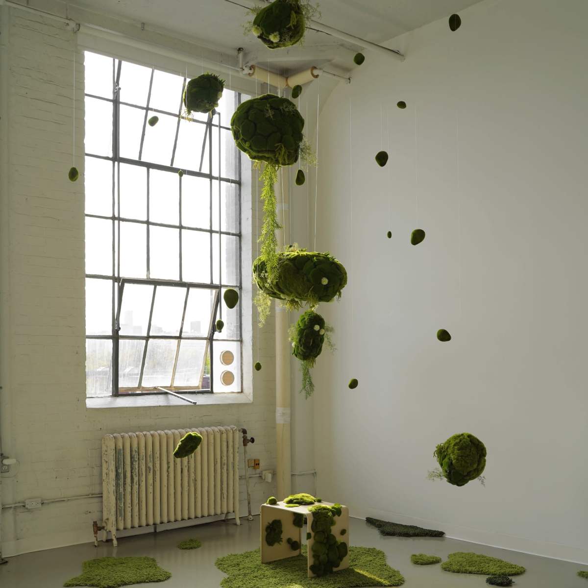 A contemporary art installation featuring green moss-like forms suspended from the ceiling and scattered on the floor within a bright, airy room.