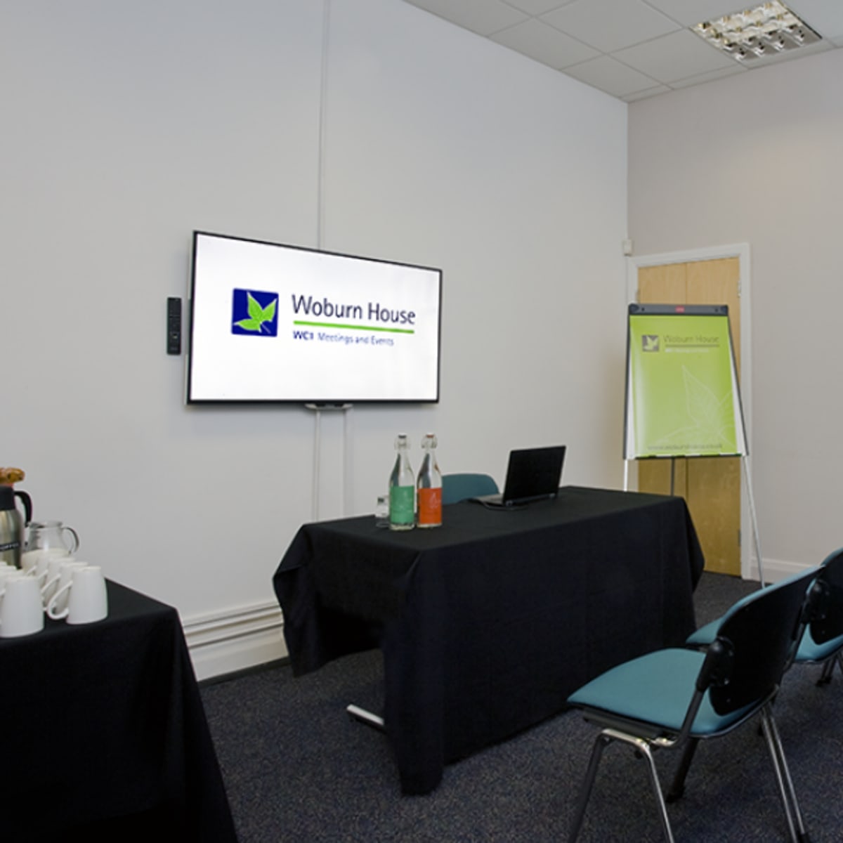 A meeting room is set up with a monitor displaying a logo, a table with beverages, and several rows of chairs.
