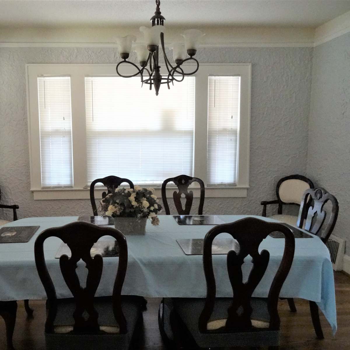 A dining room features a long table with a light blue tablecloth surrounded by six wooden chairs, with a floral centerpiece and a chandelier above.