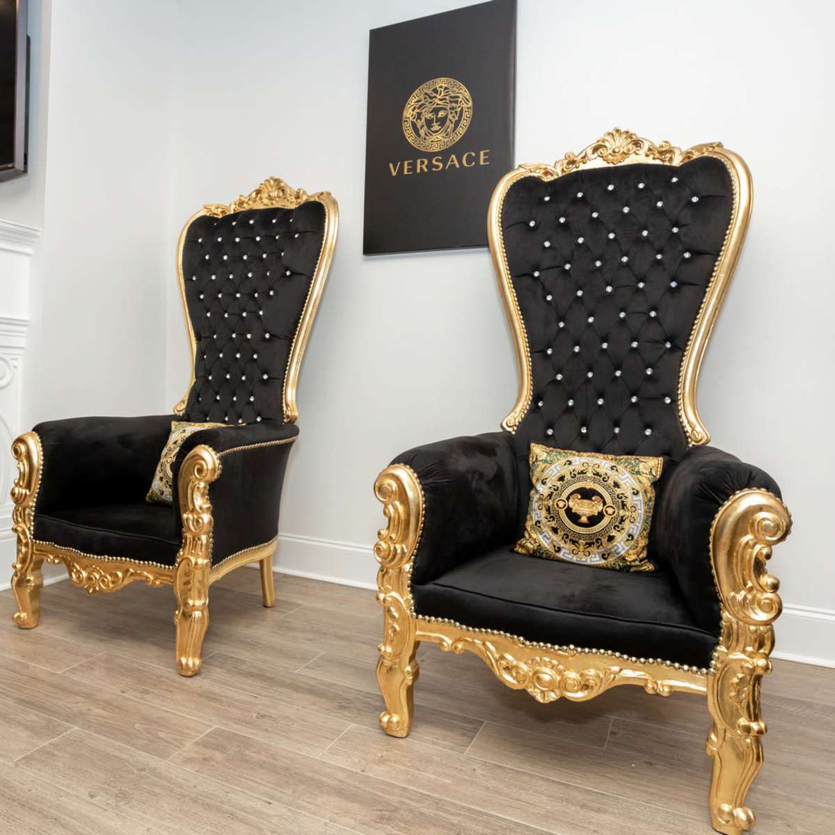 Two ornate black and gold armchairs with tufted upholstery placed next to a fireplace and a wall adorned with a Versace logo.