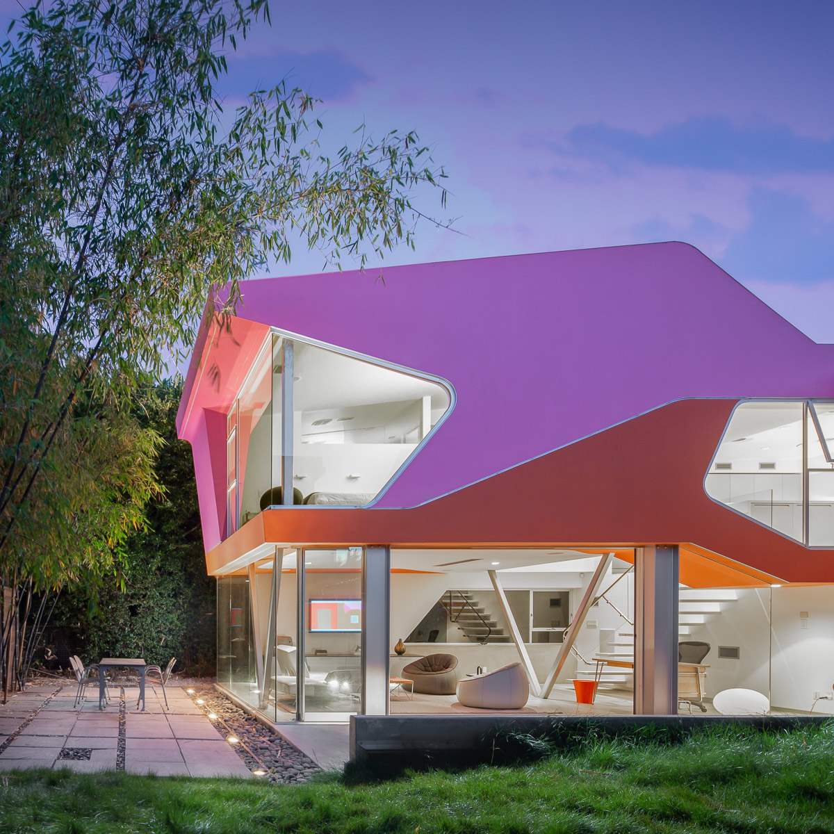 A modern house with a distinctive pink and orange exterior, featuring large glass windows and a unique angular design, surrounded by greenery and a patio area.