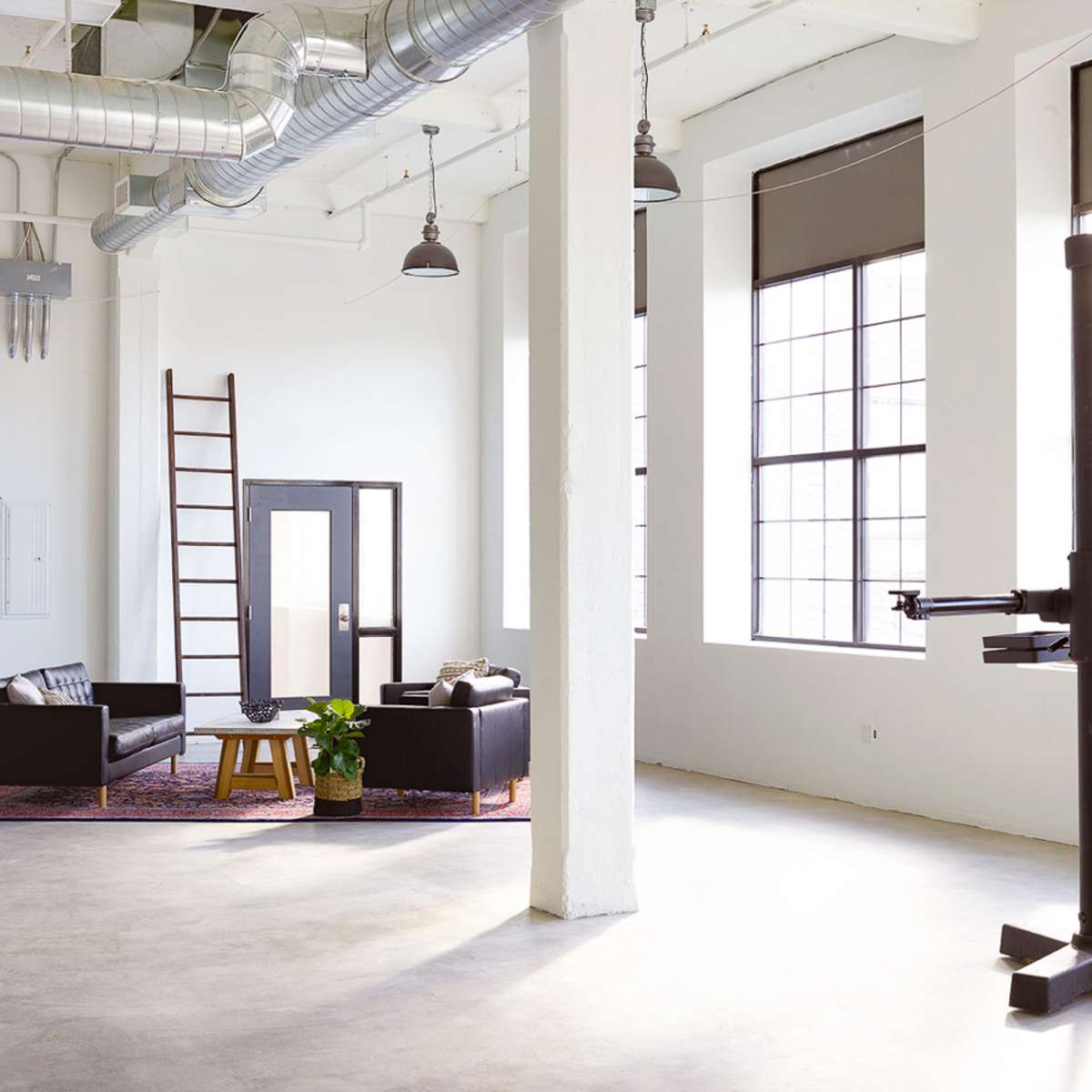 A spacious, modern interior with large windows, a ladder against the wall, and a seating area featuring black furniture and a small plant.