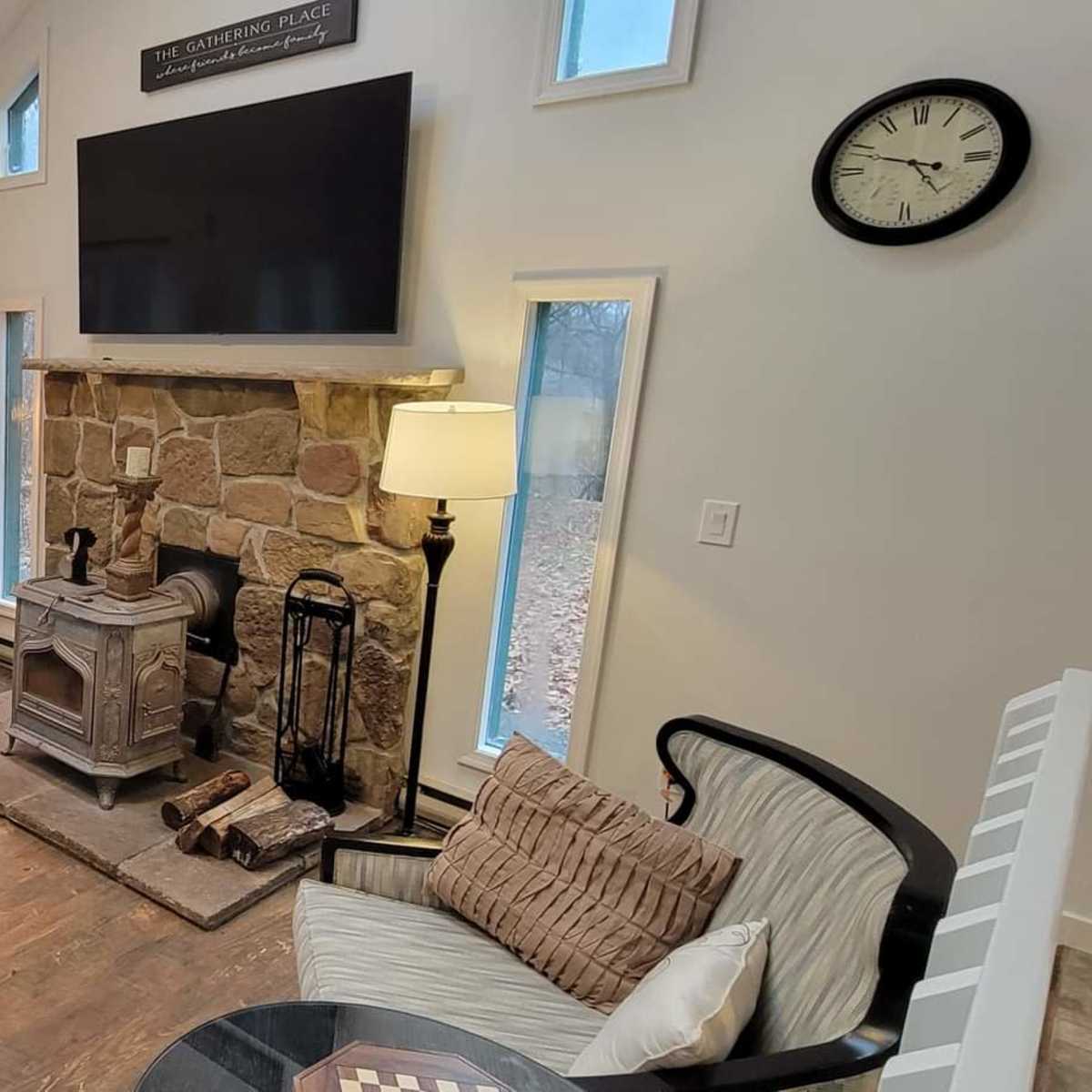 A living room with a light-colored sectional sofa, a coffee table, a stone fireplace with a wood stove, and large windows providing natural light.