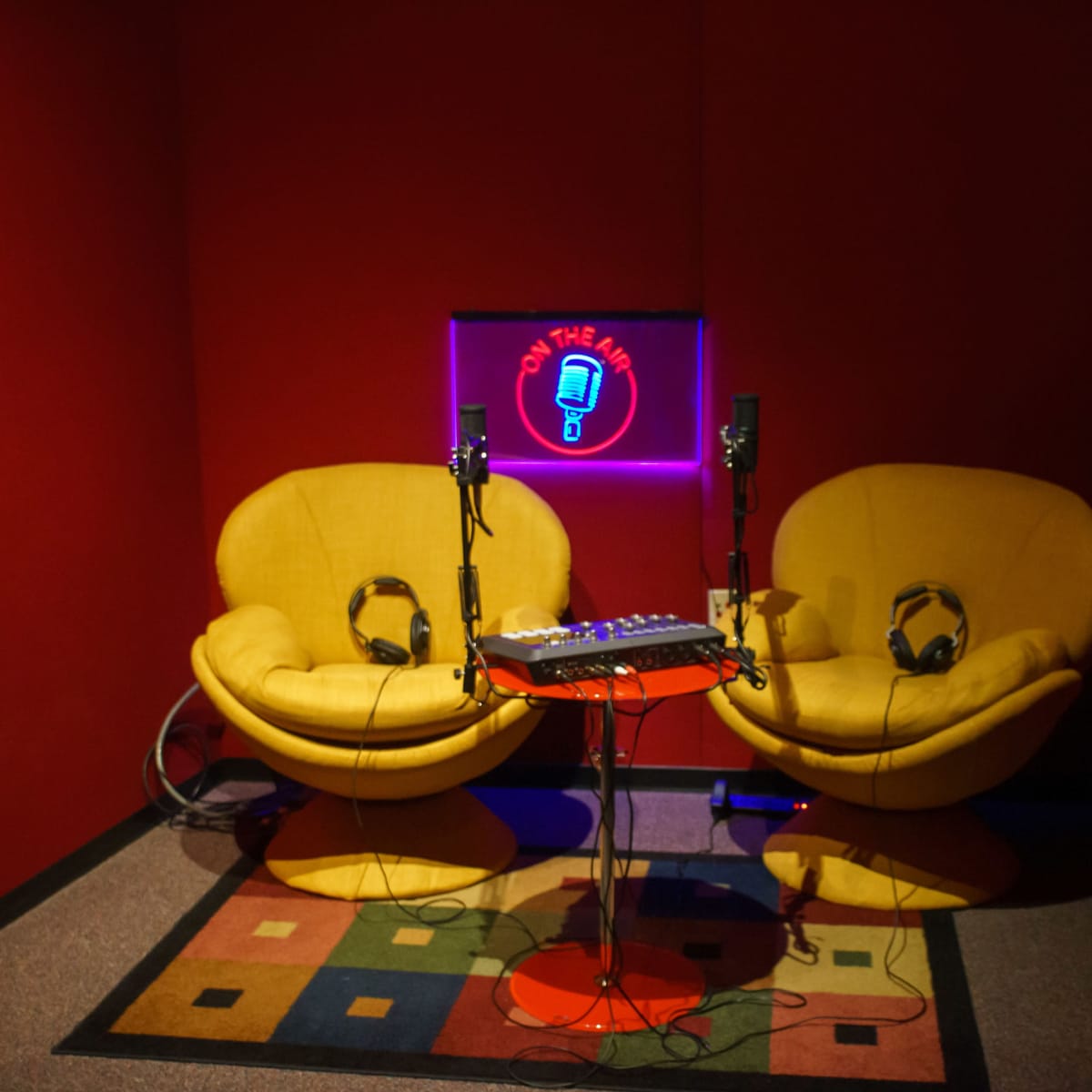 A small recording studio features two yellow chairs, microphones, and a musical mixing table against a red backdrop.