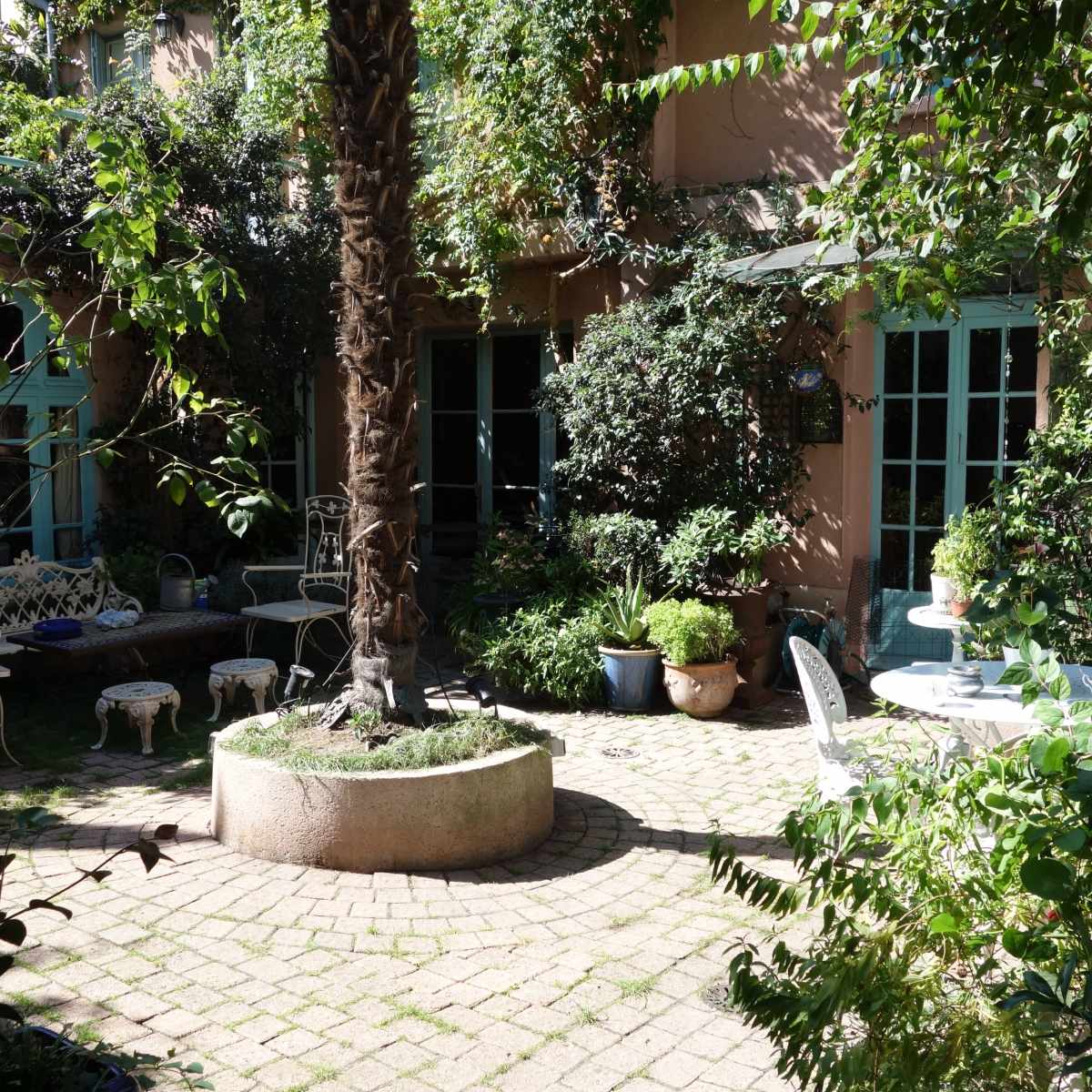 The scene features a lush courtyard with stone-paved paths, greenery, and outdoor seating arrangements surrounded by walls adorned with plants.