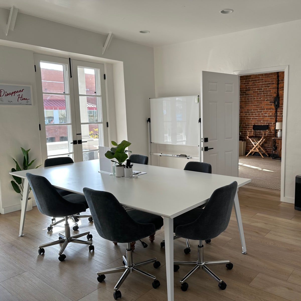 A modern office space with a large white table and gray chairs, a whiteboard, and a kitchenette displaying a small fridge and drinks.