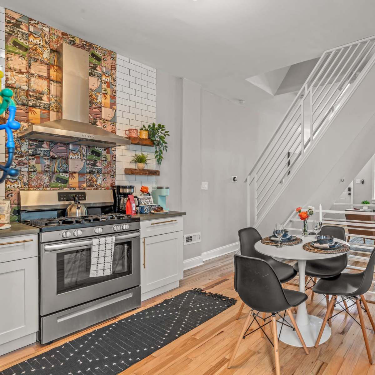 A modern kitchen area features a round dining table with black chairs, a stainless steel stove, and a tiled backsplash, leading to a living room with large windows and a sectional couch.