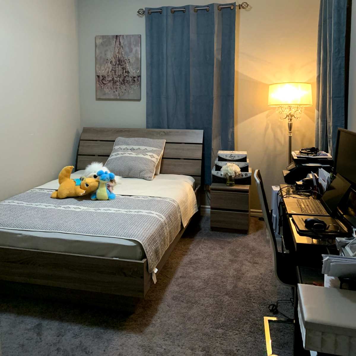 A cozy bedroom with a single bed, a nightstand, a lamp, a computer desk, and blue curtains.