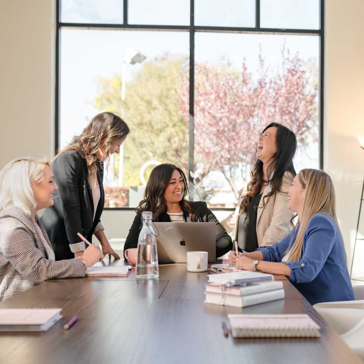 A group of five women are gathered around a table in a bright meeting room, engaged in a conversation while taking notes and using a laptop.