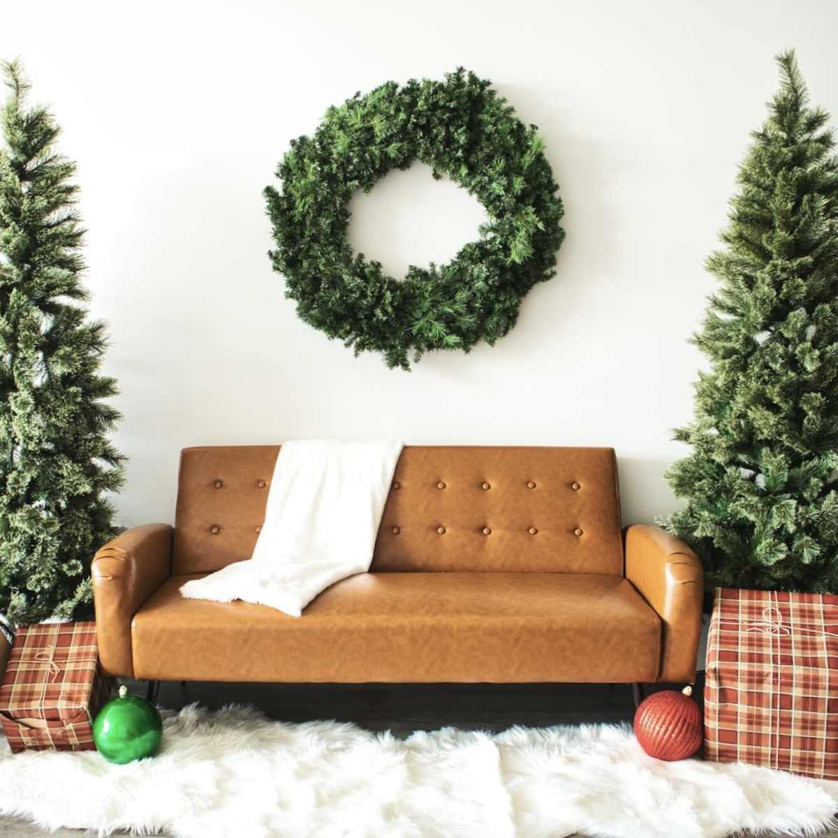 A brown leather sofa is positioned in front of two green Christmas trees and a large wreath, surrounded by wrapped gifts on a white fur rug.