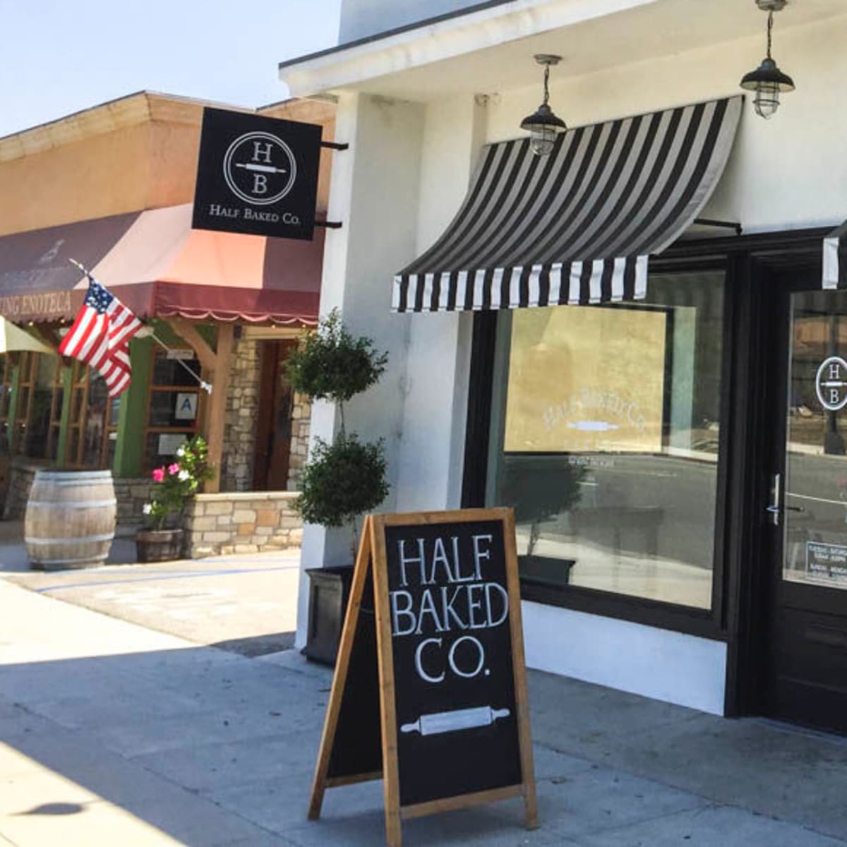 The exterior of a bakery named "Half Baked Co." features large windows with black and white striped awnings and a sidewalk sign.