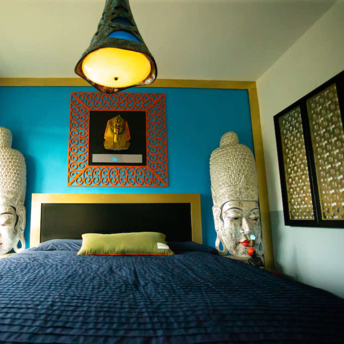 A bedroom with a blue accent wall, featuring two large Buddha sculptures on either side of a bed with a dark blue comforter and a decorative lamp hanging above.