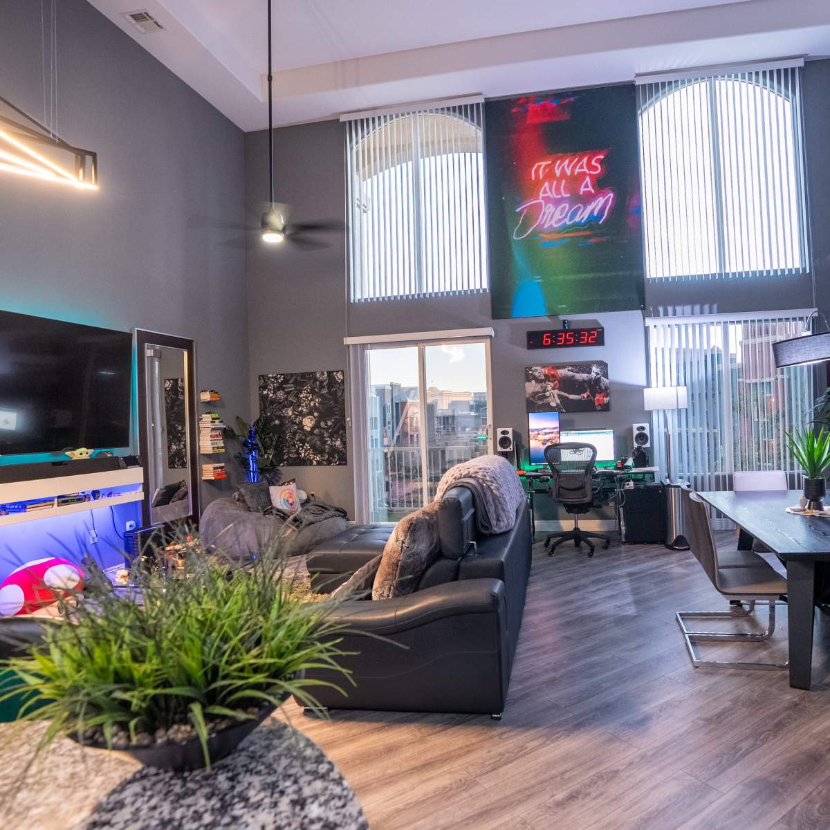 A modern living space with a high ceiling, featuring a mix of lounging and work areas, large windows, and contemporary decor including vibrant wall art and technology accessories.