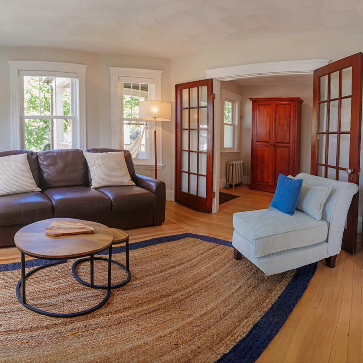 A cozy living room featuring a sofa, a light blue armchair, a circular coffee table, and large windows that allow natural light to fill the space.