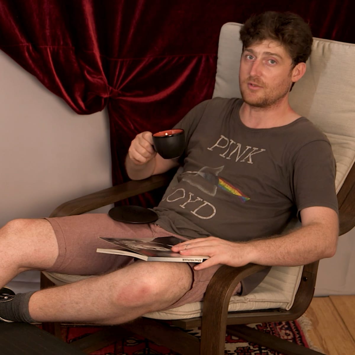 A man sits in a chair holding a cup, with a book and magazine on a side table beside him.