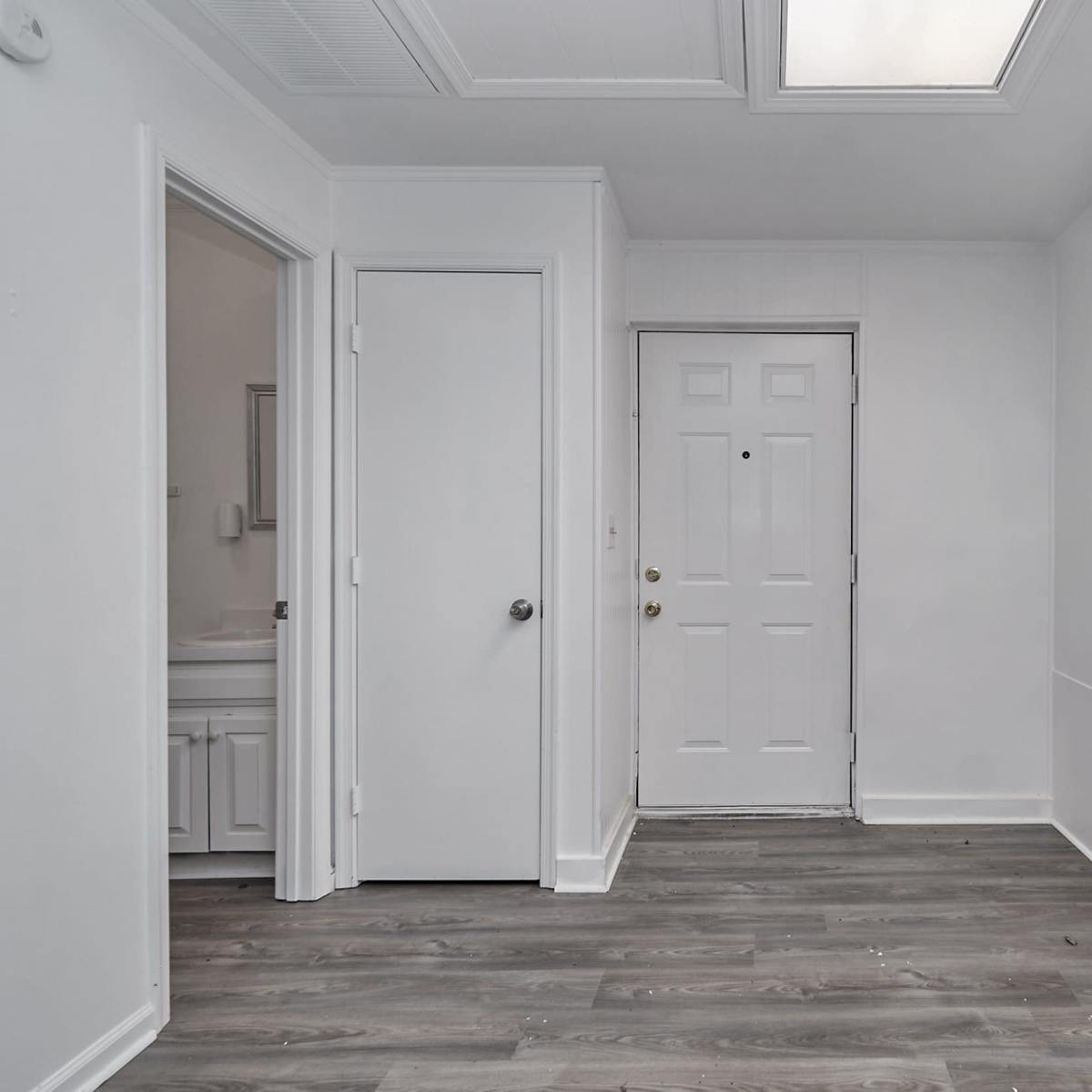 A small entryway with a front door and access to a bathroom, featuring white walls and gray laminate flooring.