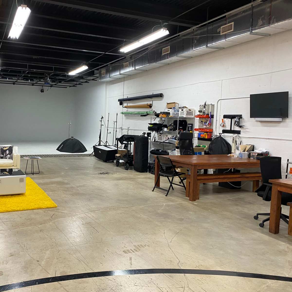 A spacious, well-lit studio featuring a minimalist seating area with a couch and yellow rug, alongside various equipment and furniture arranged throughout the concrete floor.