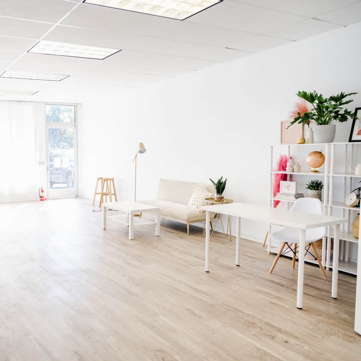 A bright, minimalist room with light-colored wooden flooring, large windows, a sofa, a desk, and decorative plants.