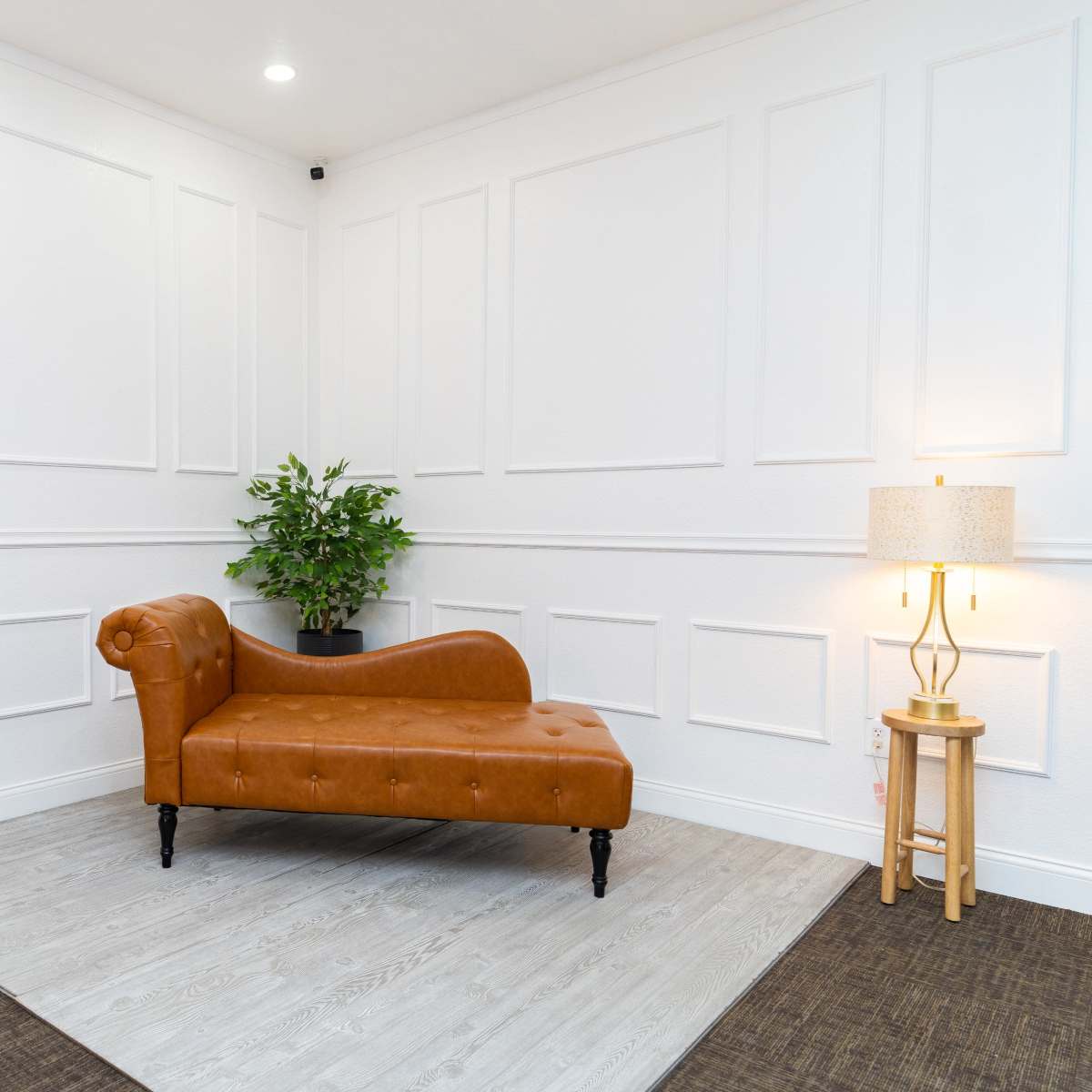 A cozy interior with a brown chaise lounge, a potted plant, and a floor lamp beside a small fireplace against a white paneled wall.
