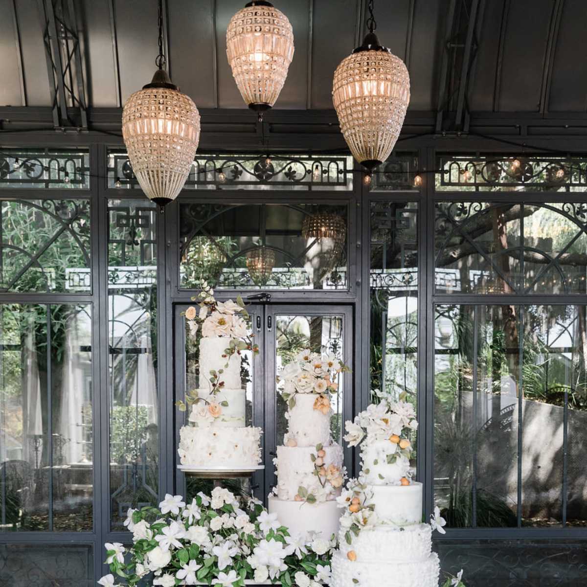 A display of several elegant white wedding cakes adorned with flowers stands on a pedestal surrounded by greenery in a beautifully designed indoor space.