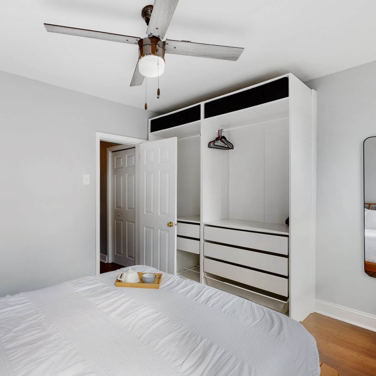 A neatly arranged bedroom features a bed with a tray on top, a ceiling fan, a closet with hanging space, and a mirror reflecting the room's interior.