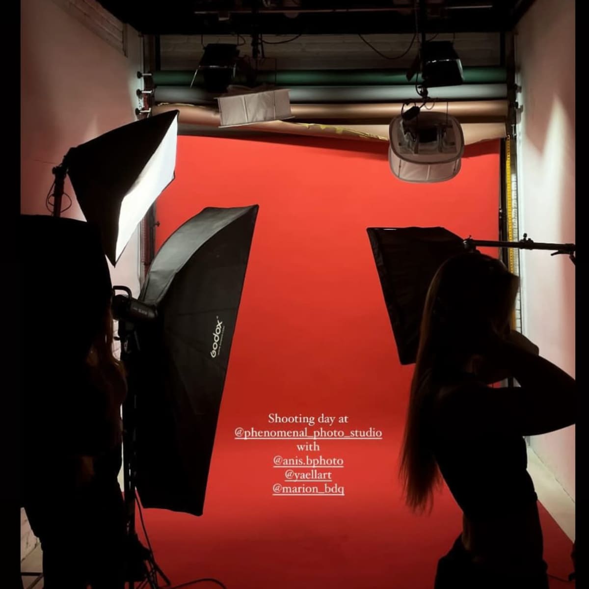 A silhouette of a model poses against a red backdrop in a photography studio, surrounded by lighting equipment.