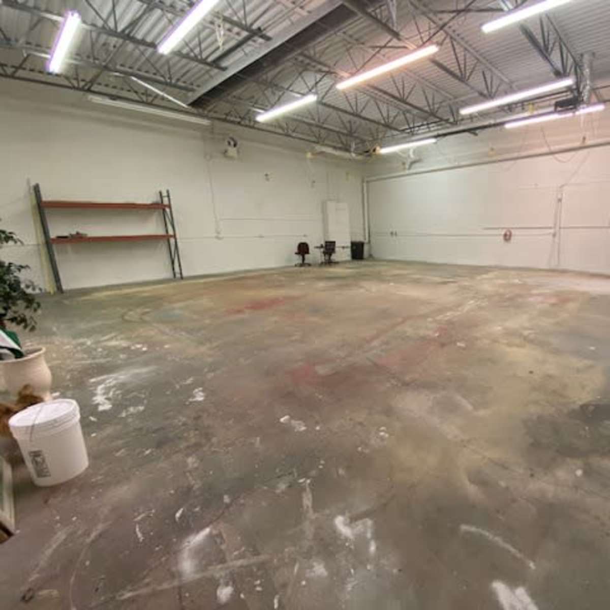 An empty industrial space with a concrete floor, metal shelving along one wall, and a staircase leading to a loft area.