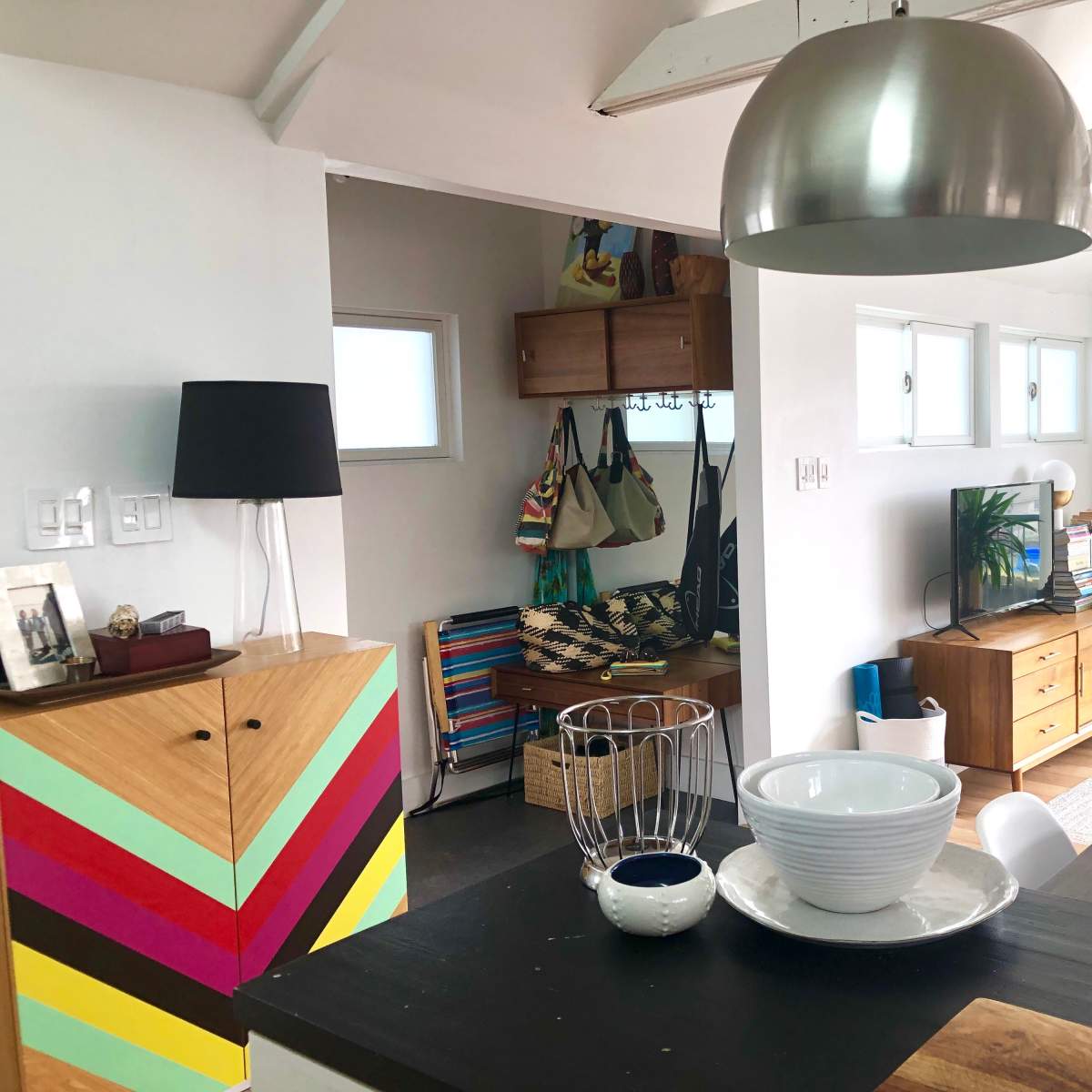 A modern living space features a dining area with a black table, colorful storage cabinet, and a well-lit corner with plants and shelves.