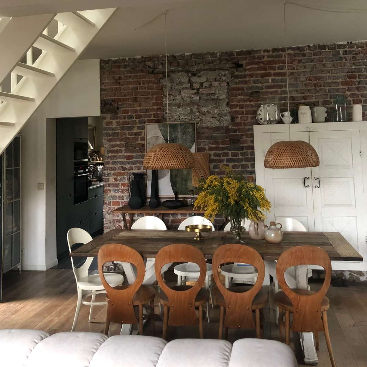 A dining area features a wooden table surrounded by chairs, with two woven pendant lights overhead and a bouquet of yellow flowers in the center.