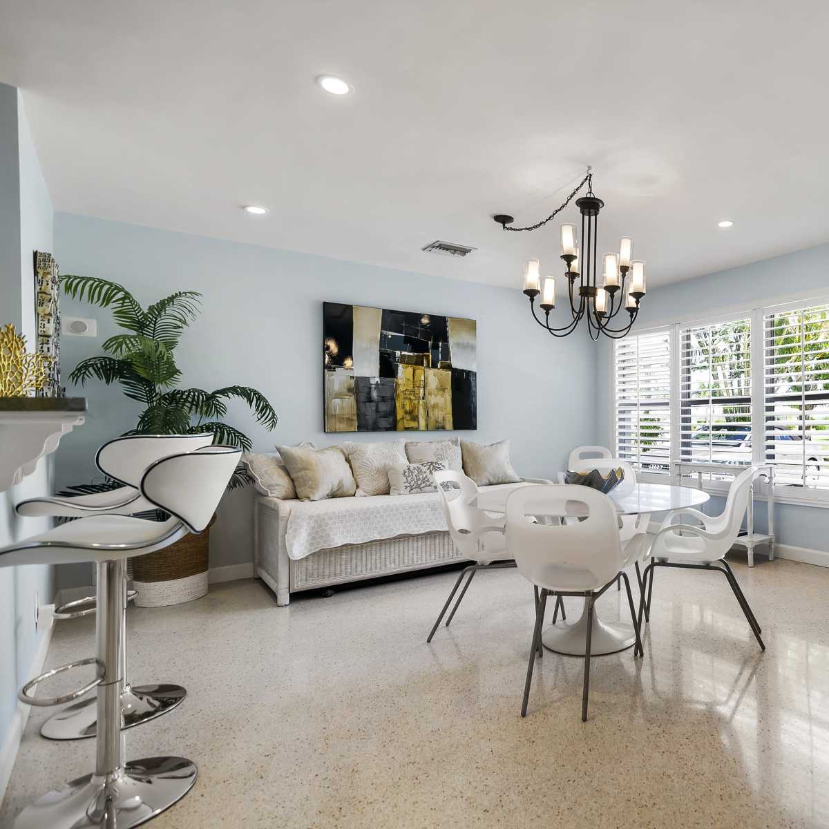A dining area features a round table with white chairs, a chandelier, a gray sofa, and a large window with shutters.