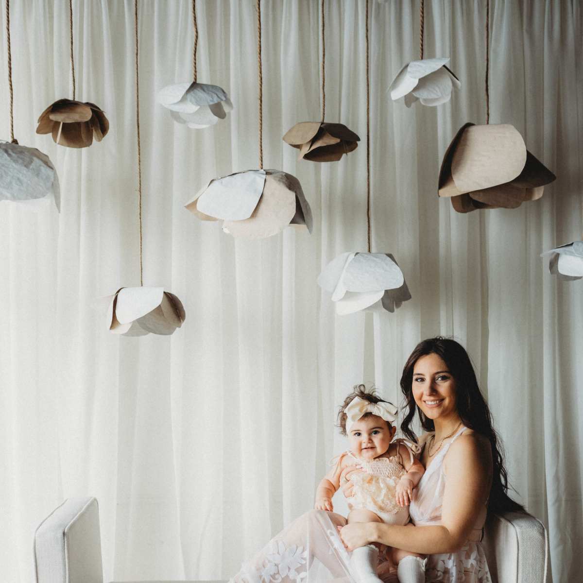 A woman in a light dress sits on a sofa holding a baby, surrounded by hanging paper flowers against a white backdrop.